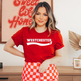 A woman wearing a red short-sleeve t-shirt with 'WESTCHESTER' printed in white across the chest, paired with red and white checkered shorts, standing in a decorated indoor setting.
