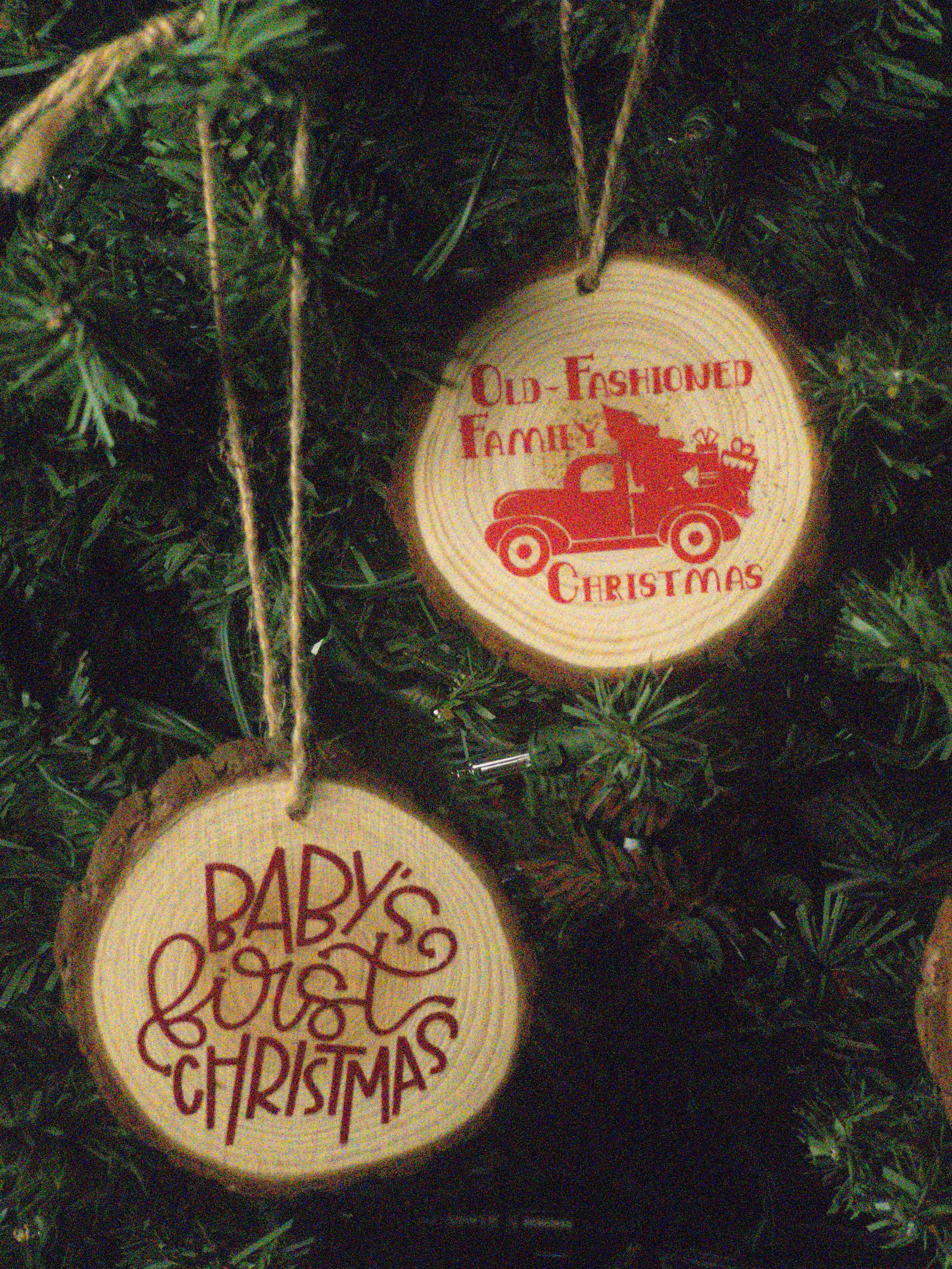 Two round wooden Christmas tree ornaments hanging on a dark green Christmas tree. One ornament is engraved with a red vintage truck and the text 'Old-Fashioned Family Christmas,' and the other is engraved with red script text reading 'Baby's First Christmas.