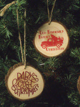 Two round wooden Christmas tree ornaments hanging on a dark green Christmas tree. One ornament is engraved with a red vintage truck and the text 'Old-Fashioned Family Christmas,' and the other is engraved with red script text reading 'Baby's First Christmas.