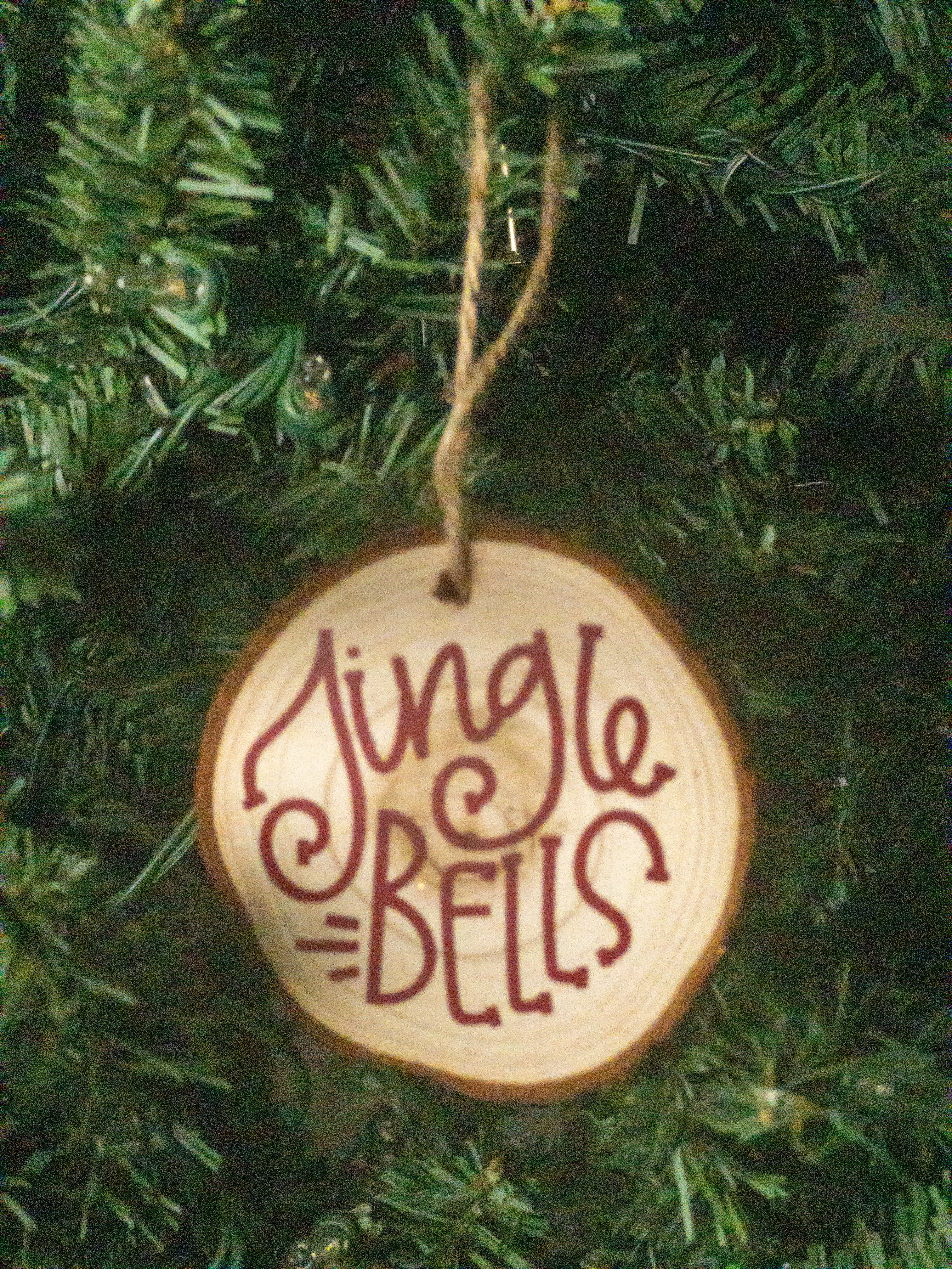 A round wooden Christmas ornament hanging from a jute string on a green Christmas tree. The ornament is made of a natural wood slice with the words 'Jingle Bells' written in dark red cursive script.