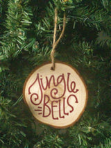 A round wooden Christmas ornament hanging from a jute string on a green Christmas tree. The ornament is made of a natural wood slice with the words 'Jingle Bells' written in dark red cursive script.