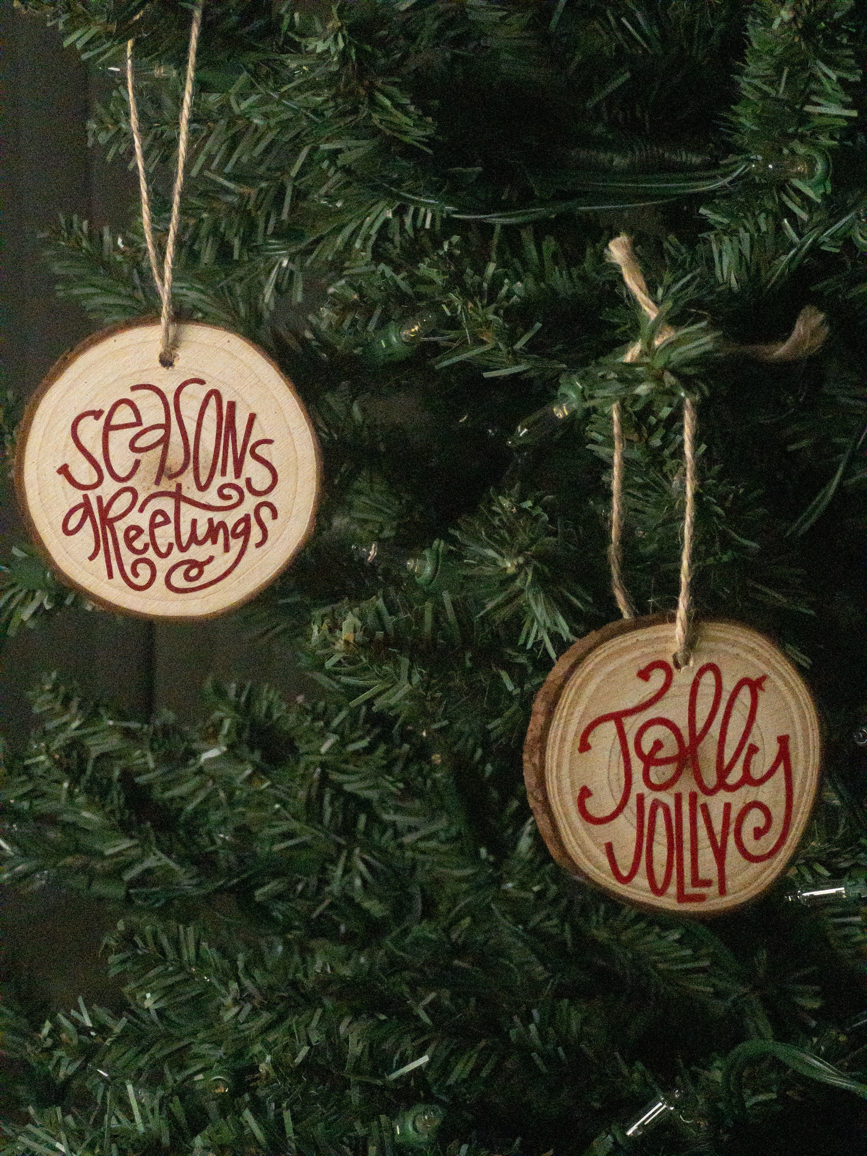 Two round wooden Christmas ornaments hanging on a green artificial tree. One ornament reads 'Seasonsreetings' in red script, and the other reads 'JollyJolly' in red script.
