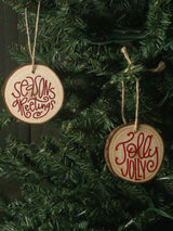 Two round wooden Christmas ornaments hanging on a green artificial tree. One ornament reads 'Seasonsreetings' in red script, and the other reads 'JollyJolly' in red script.