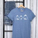 A heather blue short-sleeve t-shirt with a white graphic of three mountains and the text 'Take A Hike' across the chest, hanging on a wooden hanger next to a pair of folded blue jeans against a black and white patterned backdrop.